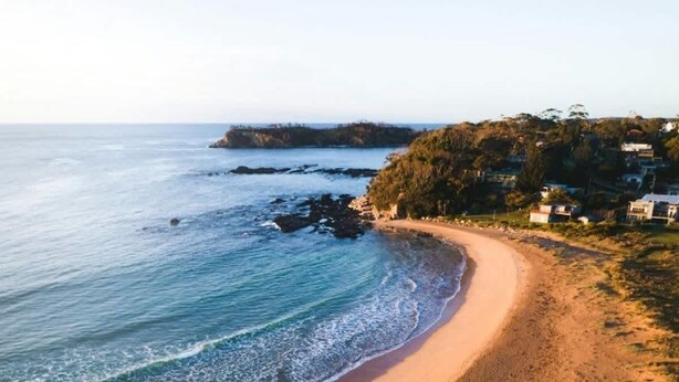 A coastal beach with a curved shoreline, a clear sky, and a distant landmass with vegetation. - Three Beaches (Malua Bay)