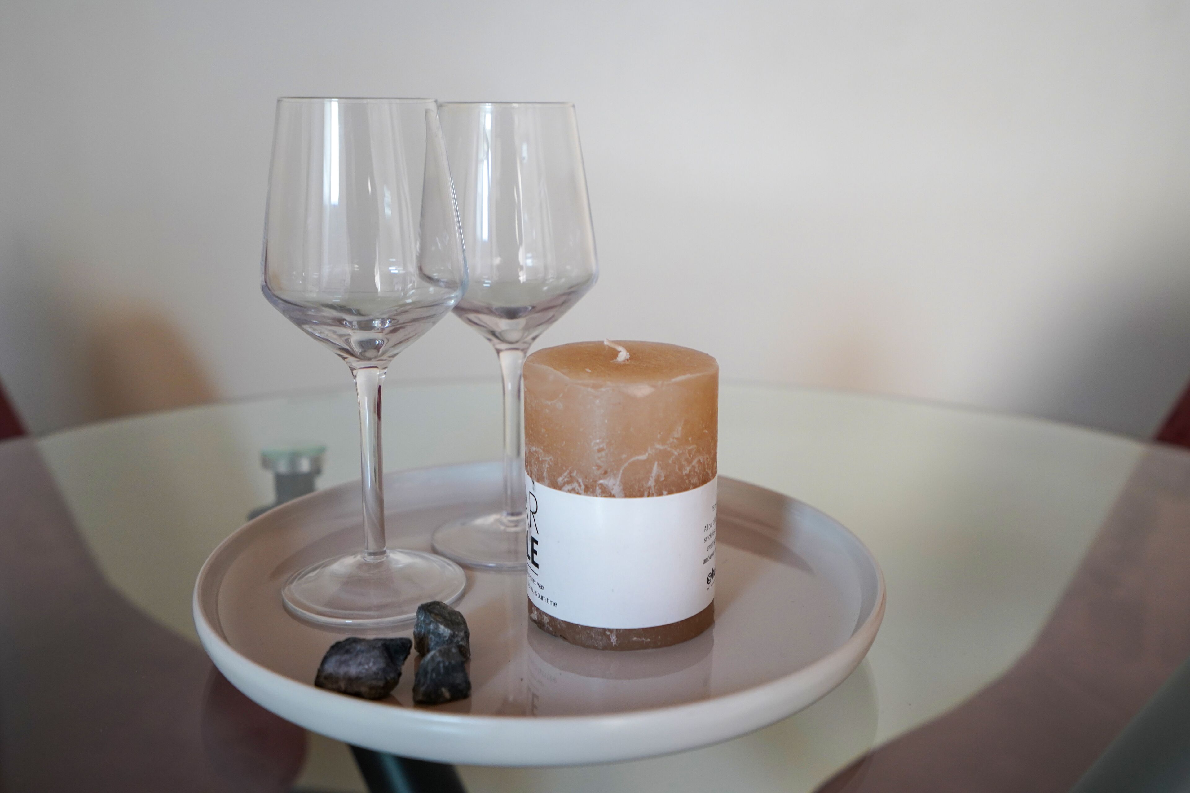 A candle with a white label on a tray, two empty glasses, and some rocks.