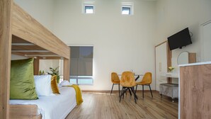 Une pièce compacte comprenant un lit superposé, une table à manger avec des chaises, un petit coin cuisine et un téléviseur fixé au mur.