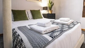 Un llit ben fet amb llençols blancs i una manta estampada, almoinos verds i una tauleta de nit amb una làmpada i una planta en test.