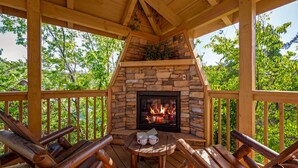 A wooden deck with a stone fireplace, surrounded by wooden furniture and a view of greenery. - Great location! Hot Tub Resort Pool Arcade, Outdoor Firepit, Free Attractions (Pigeon Forge)