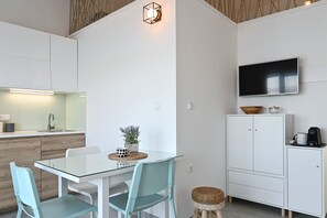 A modern kitchen with white cabinetry, a glass dining table, teal chairs, a small potted plant, and a TV mounted on the wall.