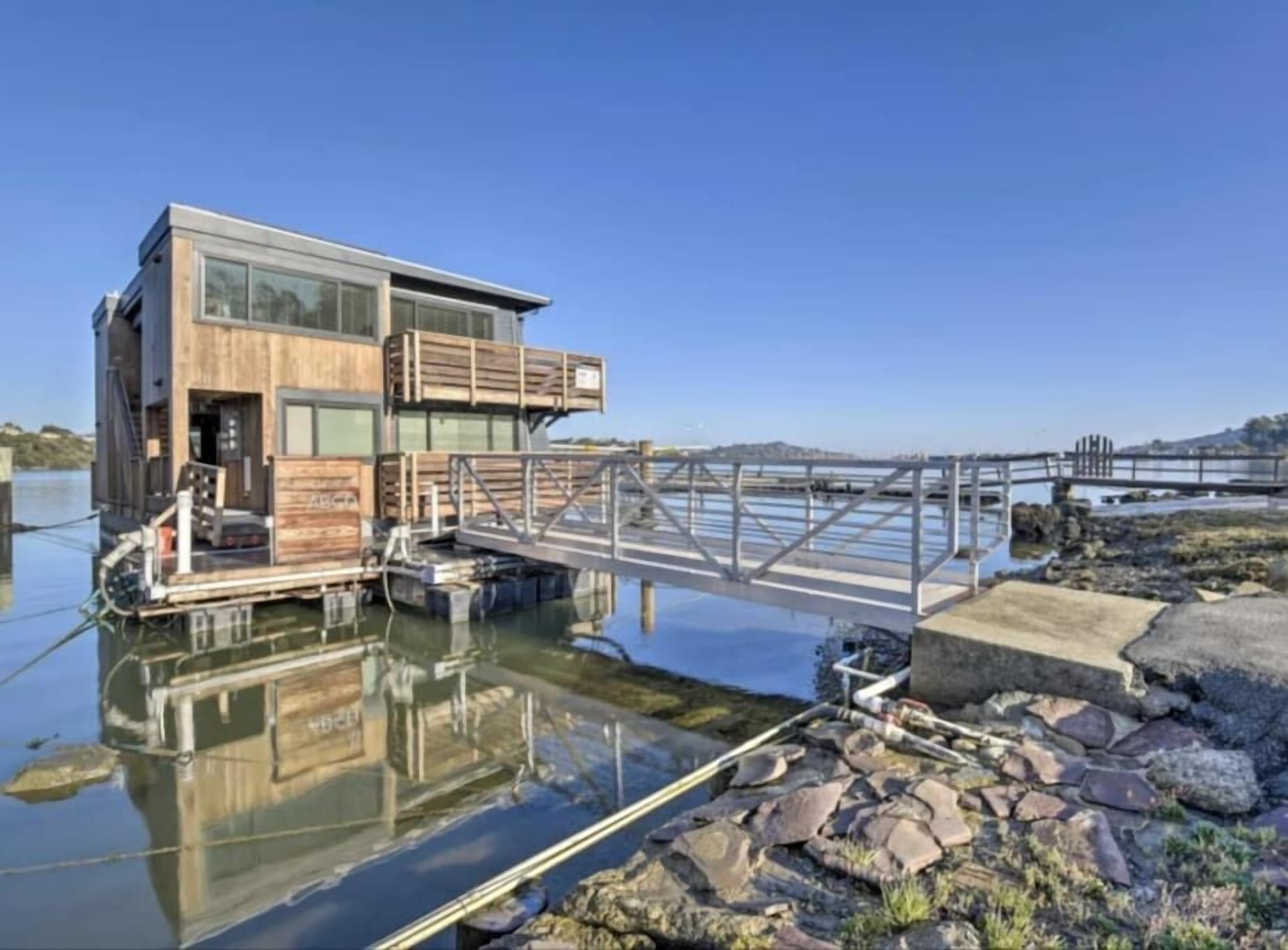 Een modern drijvend huis met een houten buitenkant en glazen ramen, verbonden met een steiger via een metalen brug.