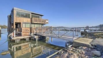 Een modern drijvend huis met een houten buitenkant en glazen ramen, verbonden met een steiger via een metalen brug.