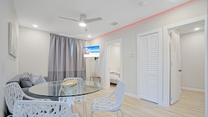 A modern living room with a glass-top dining table, white chairs, a ceiling fan, and a TV mounted on the wall. - Stylish East Little Havana Flats (Miami)