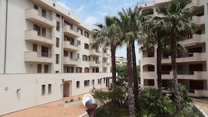 A multi-story residential building with balconies, palm trees, and a paved courtyard. - Hi Studio with beautiful sea view 🌅 Air Conditioning  (Alghero)