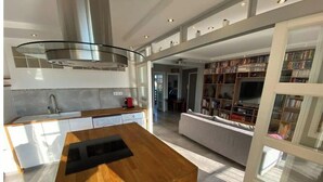 Una cocina moderna con una isla central, estufa integrada y un amplio ventanal que da a una sala de estar con sofá y estanterías.