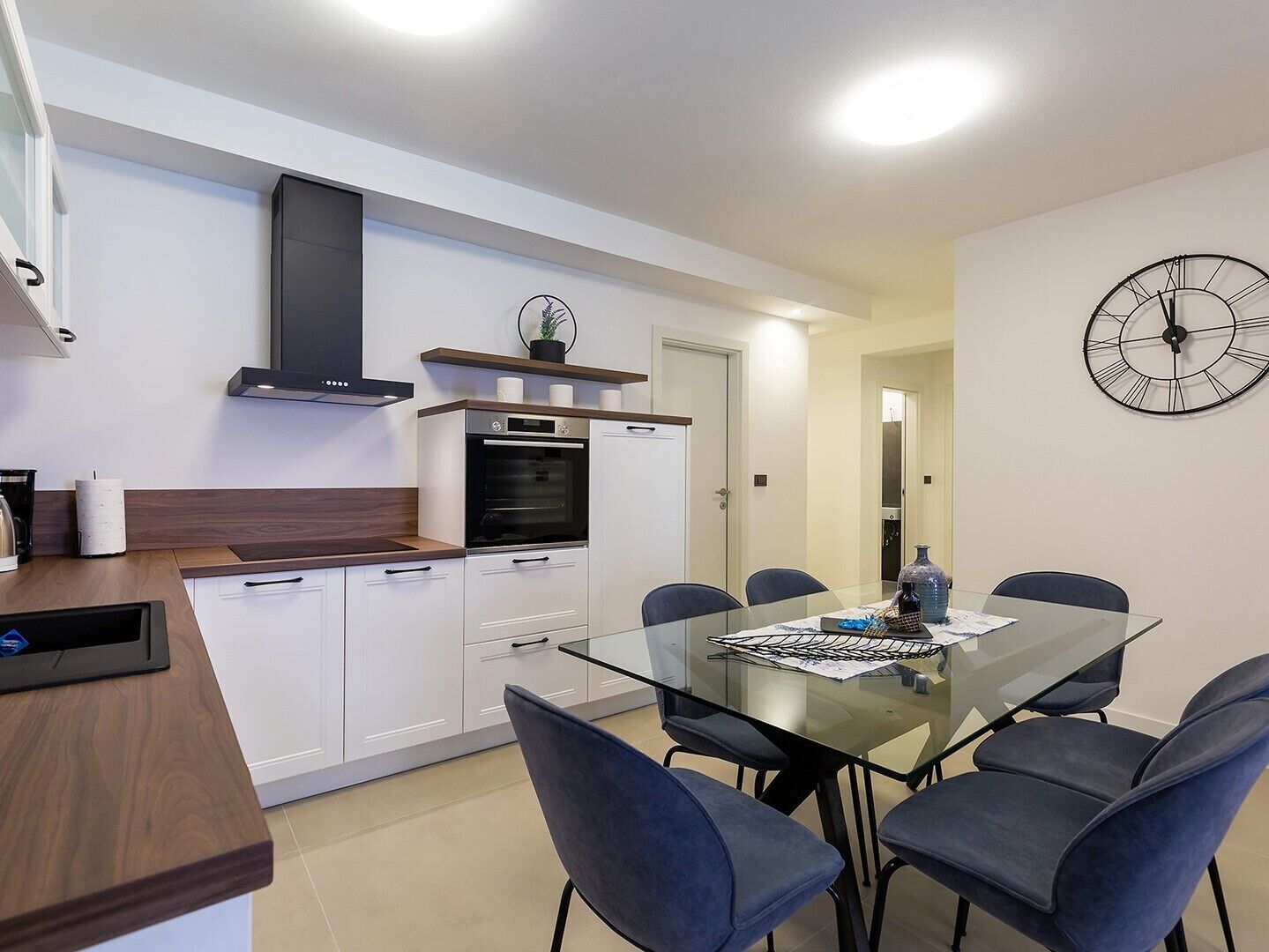 A modern kitchen with white cabinets, a built-in oven, and a glass dining table with chairs.