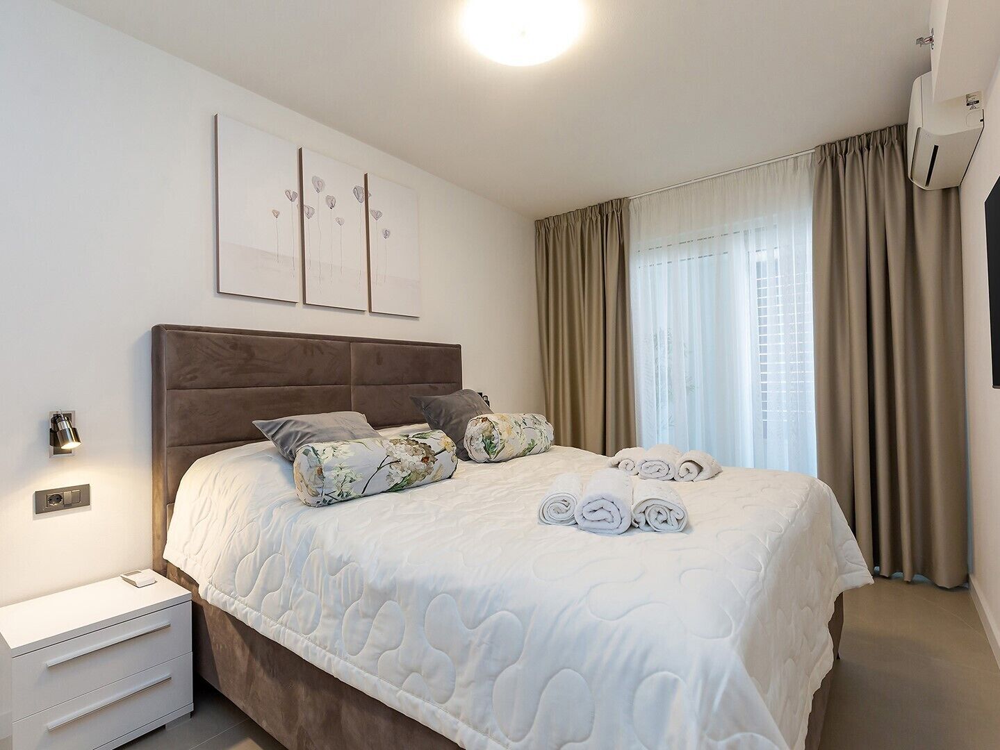 Ein ordentlich gemachtes Bett mit gesteppter Bettdecke, ein Nachttisch mit Lampe und ein Fenster mit Vorhängen.