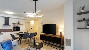 A modern living room with a sofa, a dining area with chairs, a kitchen with a stove, and a TV on a stand.