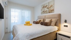 A neatly made bed with a white duvet, a set of yellow towels, and several brown pillows. There is a wall-mounted TV and a bedside table with a drawer. - Apartment with Balcony No.18 in Nar Residence (Porec)