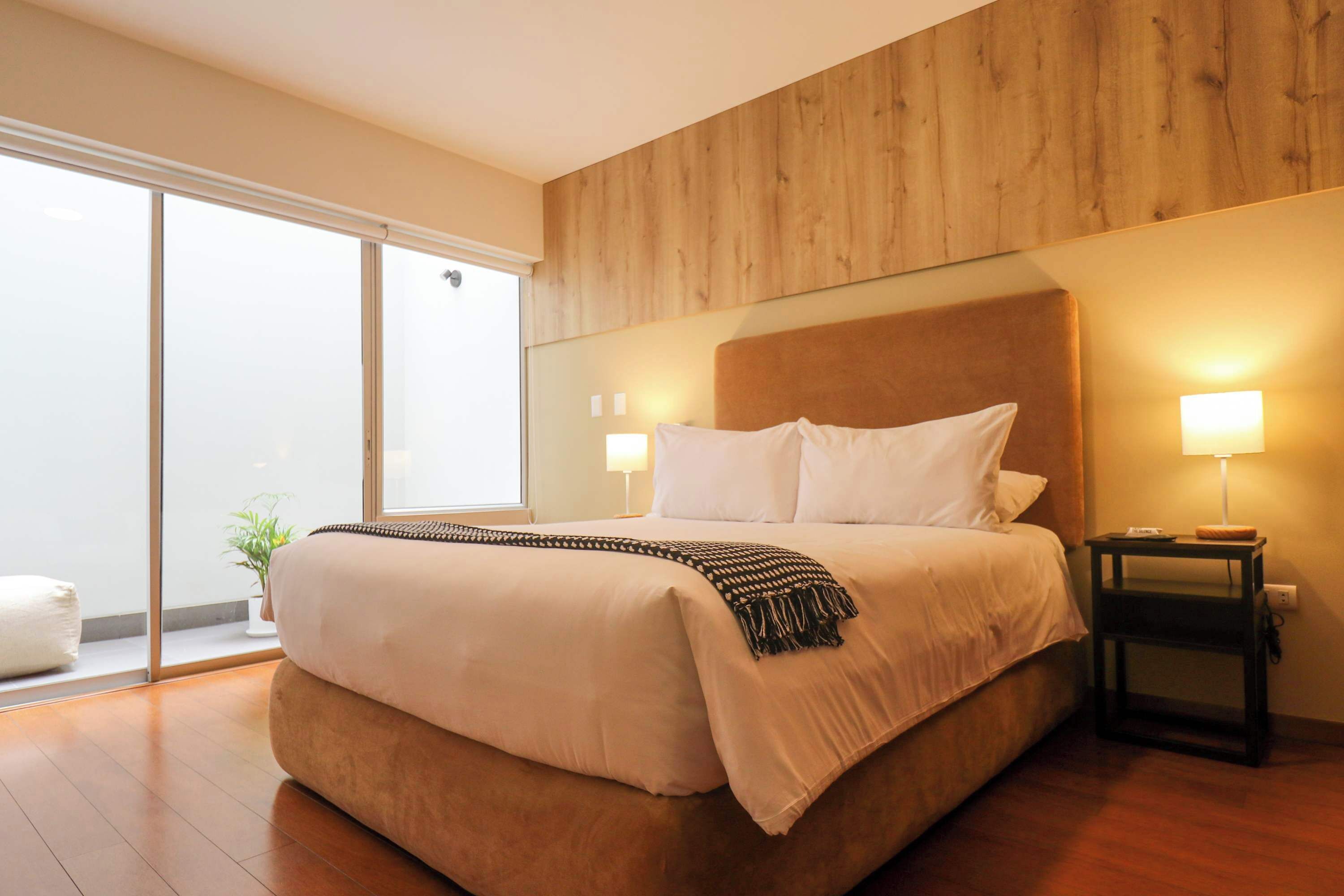 Une chambre d’hôtel moderne avec un grand lit, un mur lambrissé en bois, une porte-fenêtre coulissante et une table de chevet avec une lampe.