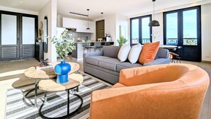A modern living room with a sofa, armchair, and coffee table, a kitchen in the background, and large windows with a view.