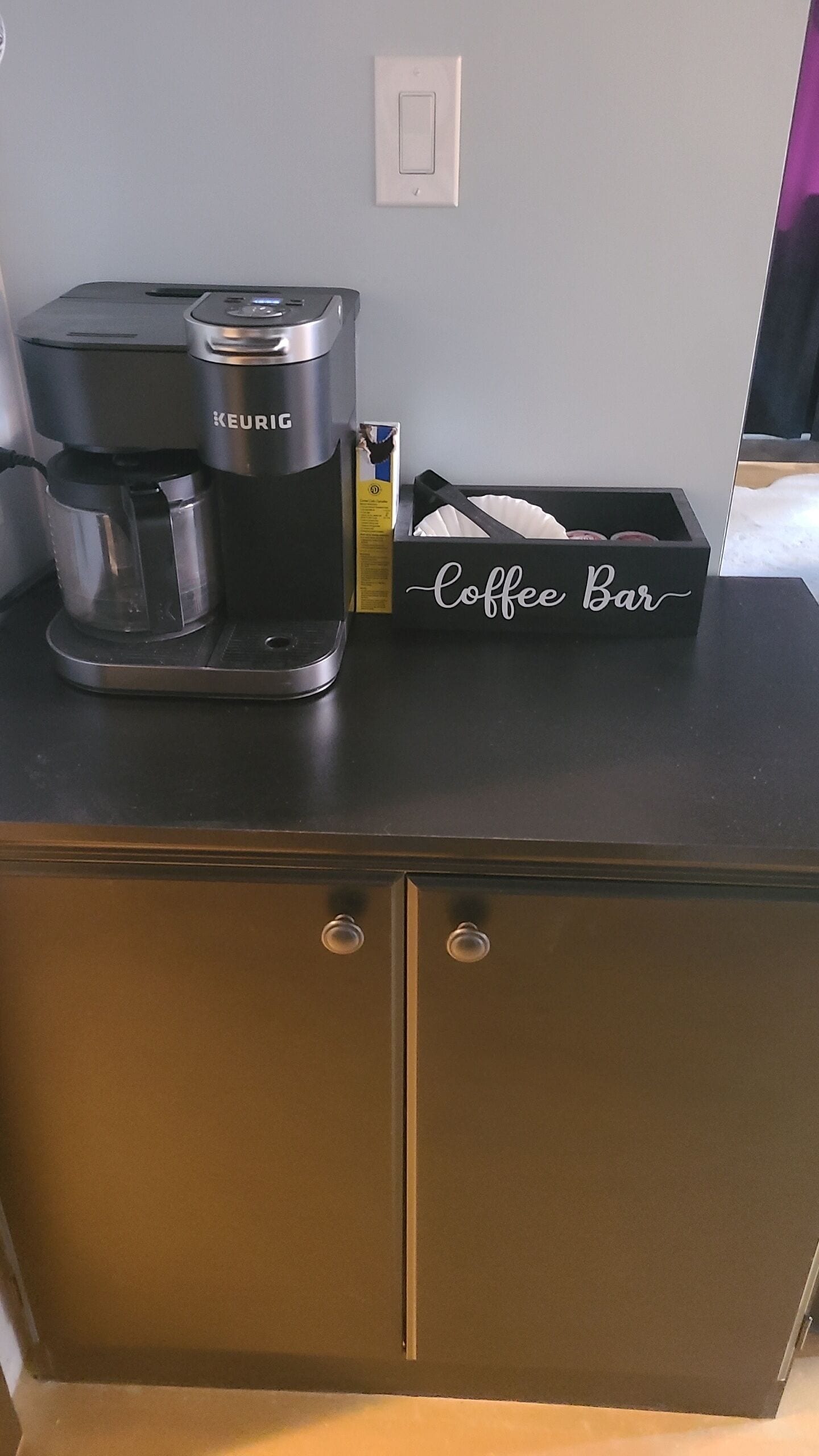 A kitchenette with a Keurig coffee maker, a coffee pod box, and a coffee bar sign on a dark countertop.