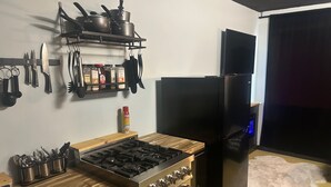 Uma cozinha compacta com fogão a gás, forno e geladeira preta. Há panelas e frigideiras em uma prateleira, e uma lata de PAM no balcão.
