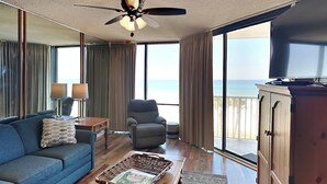 A spacious living room with a large window offering a view of the beach, a ceiling fan, a television, and a wooden cabinet. - Edgewater Windward #204 | Gulf Front + Amenities (Panama City Beach)