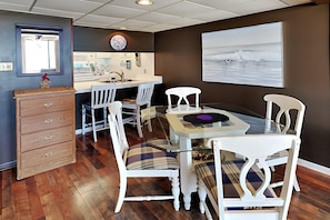 A dining area with a glass table, white chairs, a wooden dresser, and a kitchen in the background.