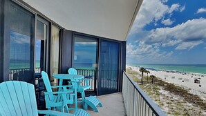 Un balcón con sillones Adirondack turquesa y una mesa, con vista a una playa con sombrillas y palmeras.
