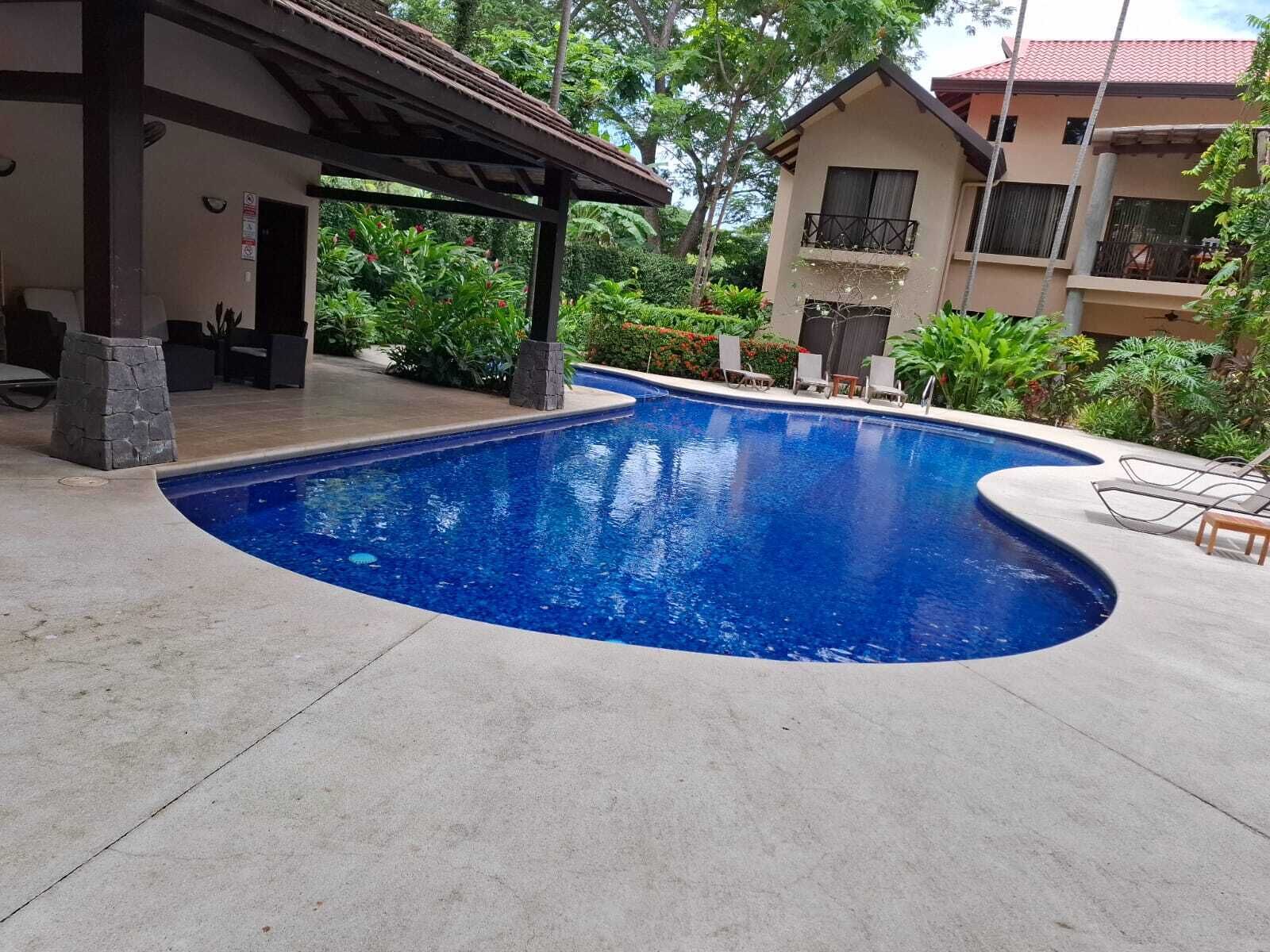 Une piscine à bord incurvé, entourée d’un patio couvert doté de piliers en pierre et de mobilier d’extérieur.
