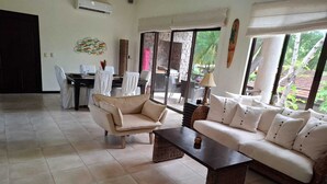 A spacious living area with a sofa, armchair, and coffee table. There is a dining table with chairs, a stone fireplace, and large windows with a view of greenery.