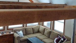 A loft-style living room with a high ceiling, exposed wooden beams, a ceiling fan, and a large window providing natural light. - White Tail Cabin located on 100 private forested acres, in a Crown Game Preserve (Apsley)