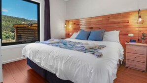 A bedroom with a large bed, wooden headboard, a nightstand, and a window with a view of greenery. - Alpine Twin Heights (Merrijig)