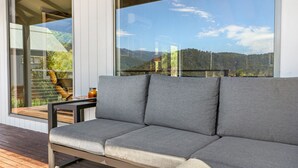 A grey sofa on a wooden deck with a large window offering a view of a mountainous landscape. - Alpine Twin Heights (Merrijig)