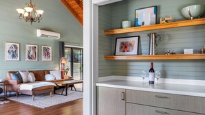 A modern living room with a brown leather sofa, a wooden coffee table, and a chandelier. There is a kitchen area with wooden shelves, a framed painting, and a wine bottle. - Alpine Twin Heights (Merrijig)