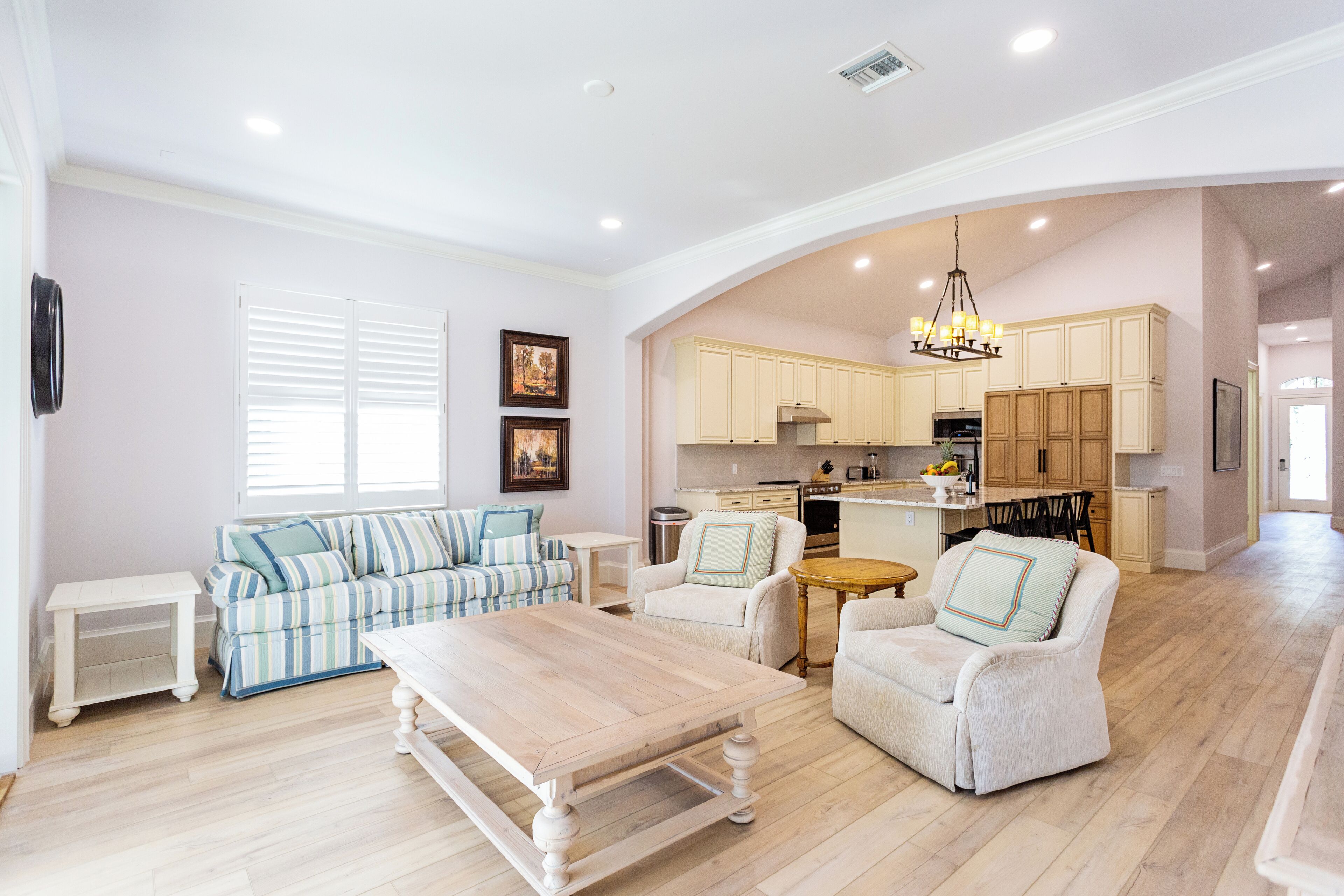 A spacious living area with a sofa, chairs, and a coffee table. A kitchen is visible in the background with wooden cabinets and a chandelier.