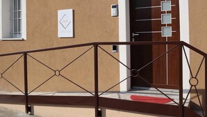 A building facade with a brown textured wall, a white door with a geometric design, a small window with metal bars, and a metal railing in front.