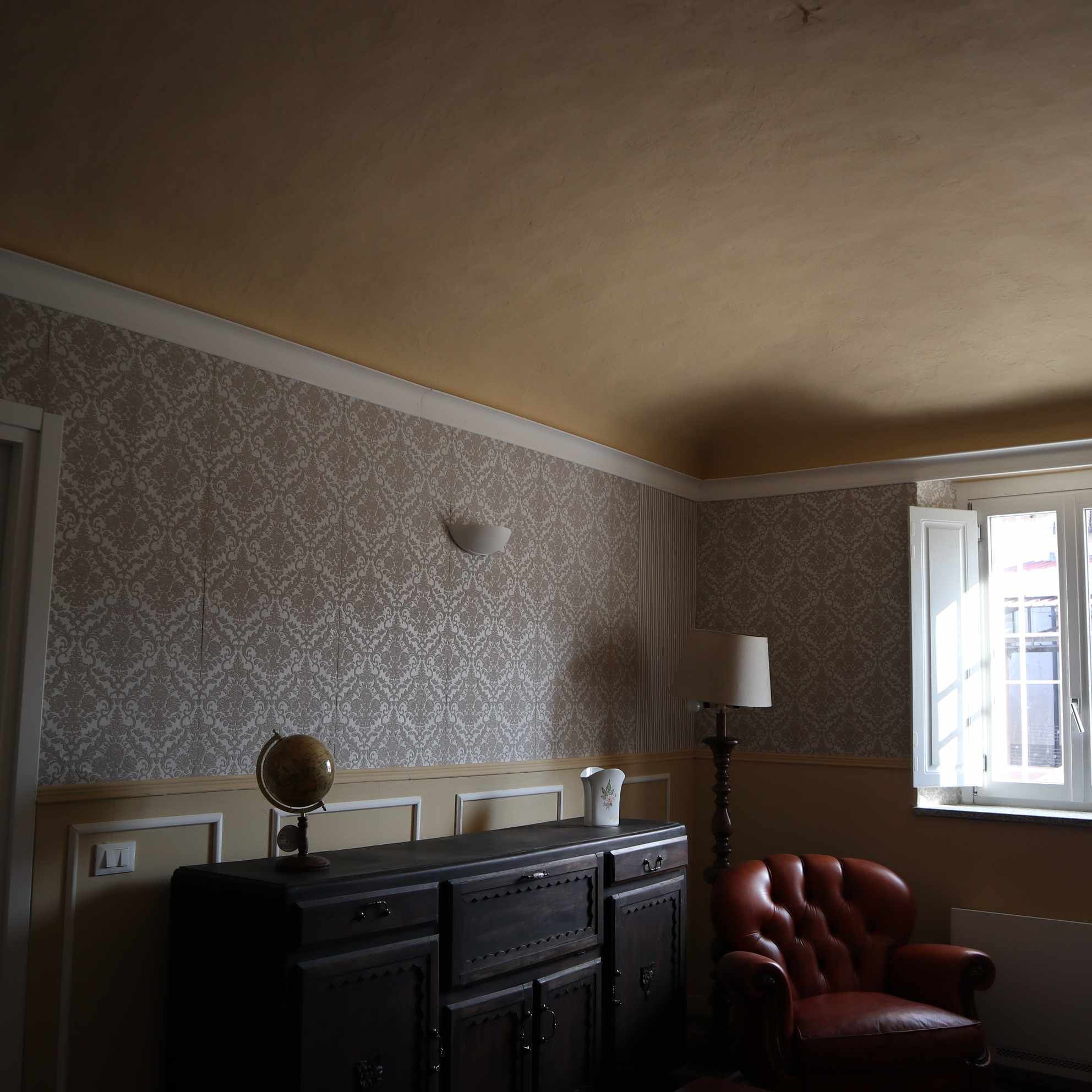 A room with patterned wallpaper, a dark wooden cabinet, a red leather chair, and a window with a view of buildings.