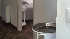 A narrow hallway with a white radiator, a small round table with a glass top, and a bed with a pillow and a painting on the wall.