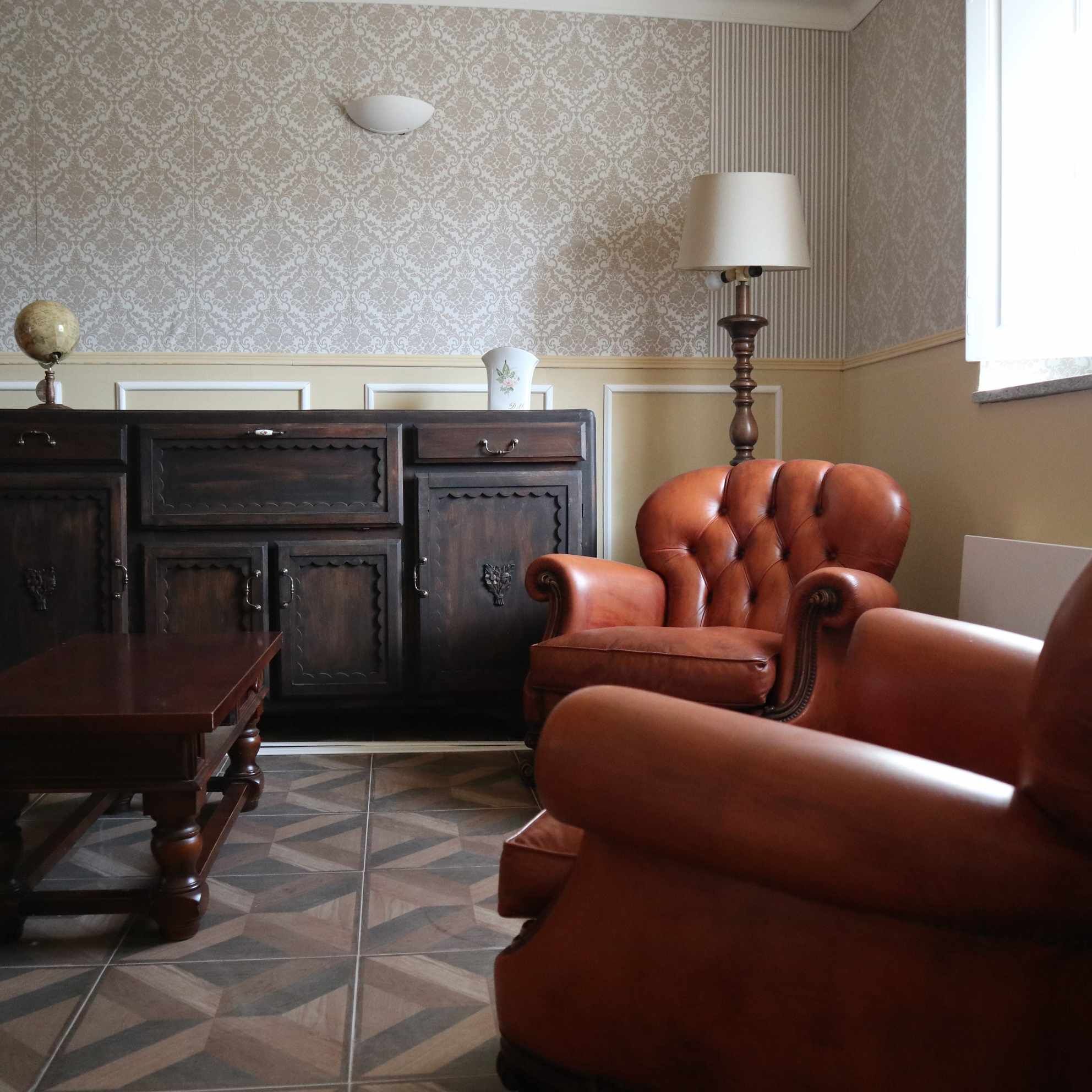 Um quarto com papel de parede estampado, um armário de madeira escura, uma poltrona de couro, uma mesa de madeira e um chão com padrão geométrico de azulejos.