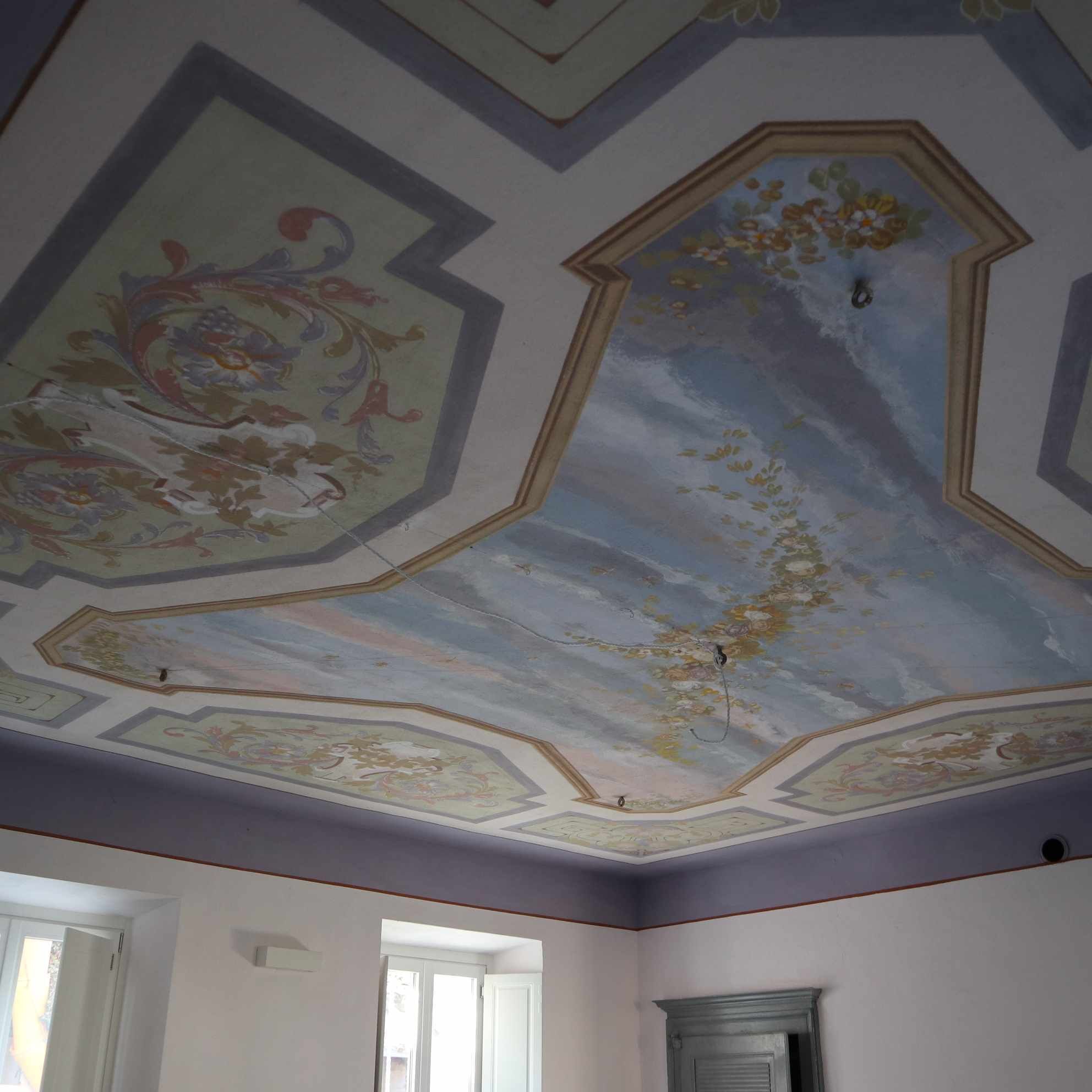 A room with a ceiling featuring ornate floral and geometric patterns, white walls, and a window with white shutters.