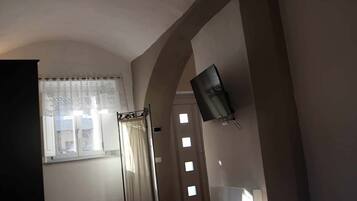 Una habitación con techo inclinado, una cama, un televisor montado en la pared y un ventanal con cortinas.