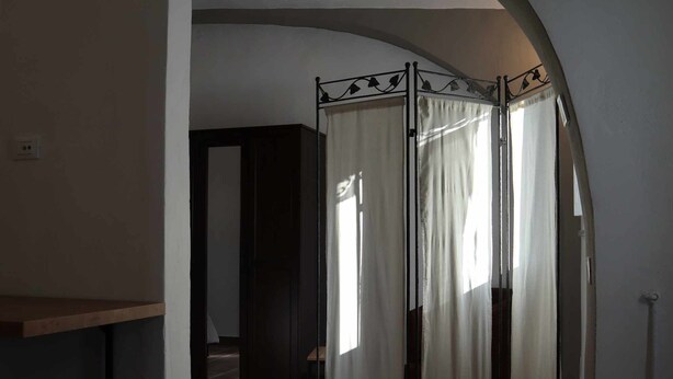 Une pièce dotée d’une cloison avec une structure métallique et des rideaux blancs, d’un sol carrelé et d’une porte-fenêtre donnant sur une autre pièce.
