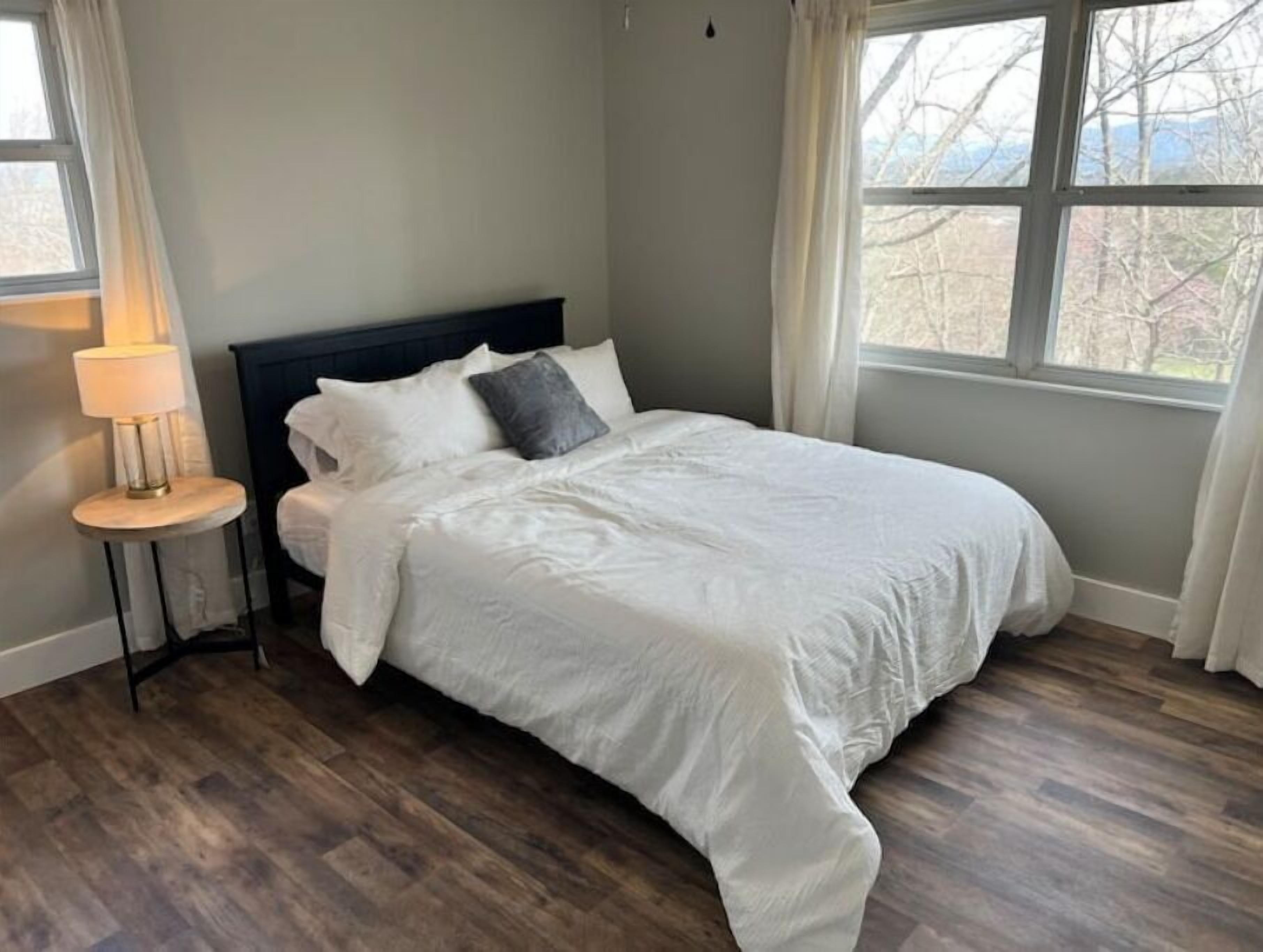 Un dormitorio con cama, mesita de noche, lámpara y ventana con vista a árboles.
