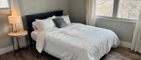 Un dormitorio con cama, mesita de noche, lámpara y ventana con vista a árboles.