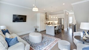 Een moderne woonkamer met een bank, voetenbankje en een getextureerd vloerkleed. In de achtergrond is een keuken met witte kasten en een centraal keukenblok zichtbaar.