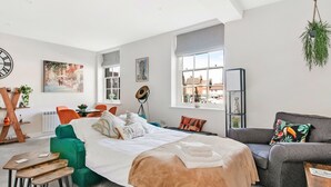 Ein modernes Schlafzimmer mit einem großen Bett, einem grauen Sofa und einem grünen Sessel. Ein Fenster bietet Blick auf die äußere Bebauung.