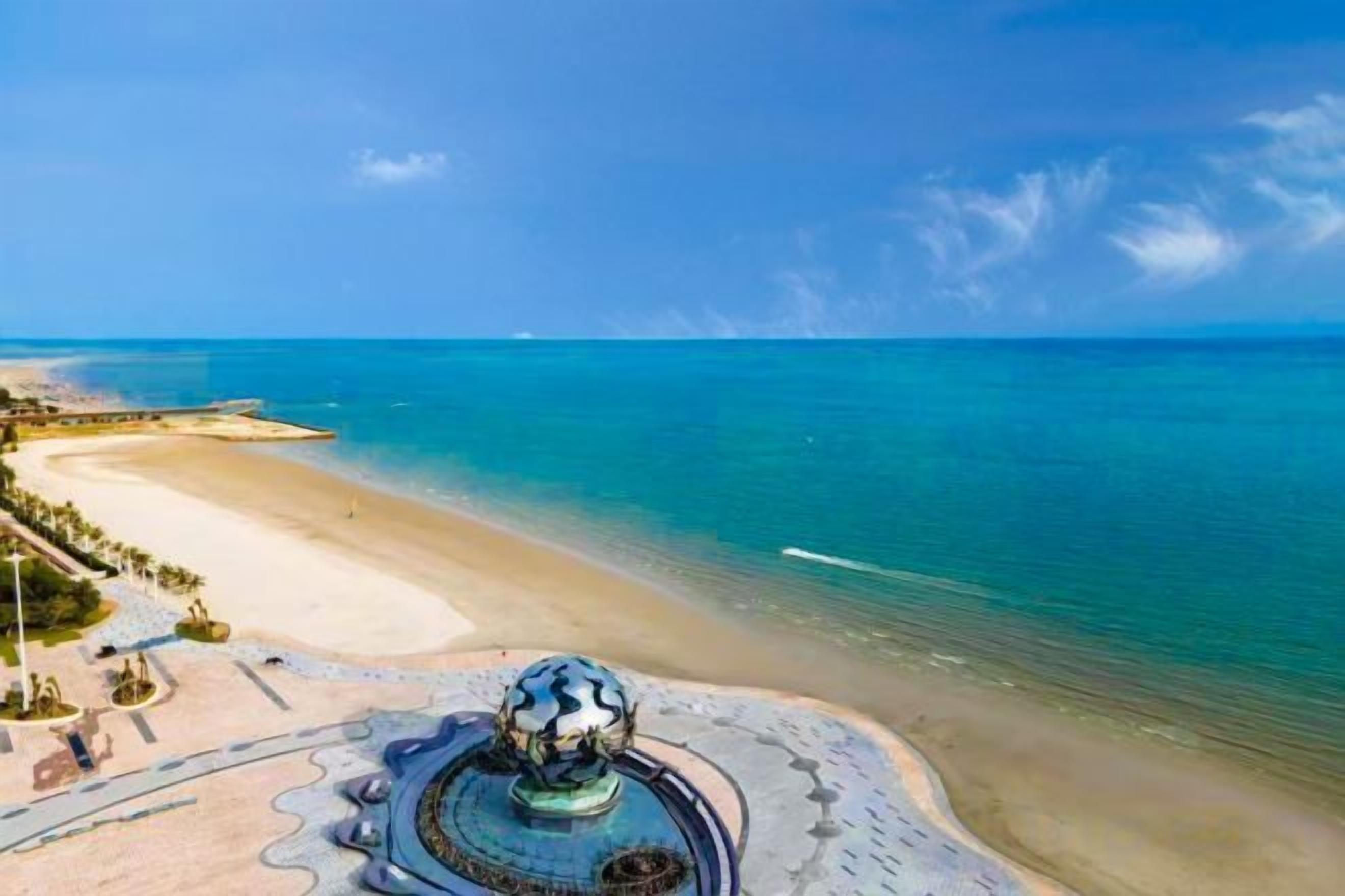 A coastal view with a beach, a fountain, and a clear sky.