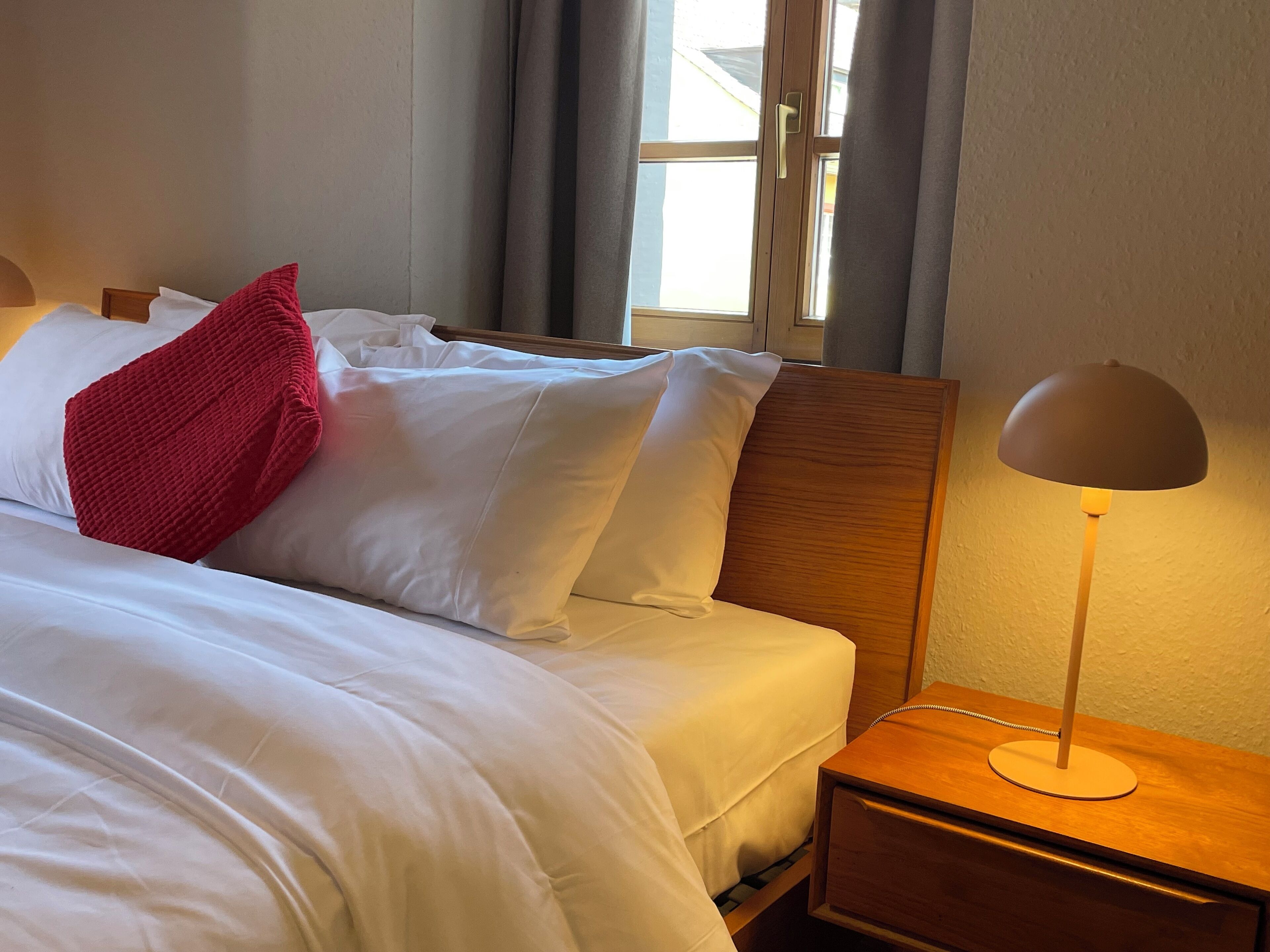Un lit bien fait, avec du linge de lit blanc et un coussin rouge, une tête de lit en bois, une lampe de chevet et une fenêtre avec des rideaux.