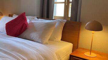 Un lit bien fait, avec du linge de lit blanc et un coussin rouge, une tête de lit en bois, une lampe de chevet et une fenêtre avec des rideaux.