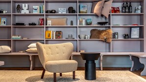 A modern living room with a beige armchair, a black coffee table, and a bookshelf filled with books, decorative items, and a small sign. - Hotel Deville Staufen (Staufen im Breisgau)