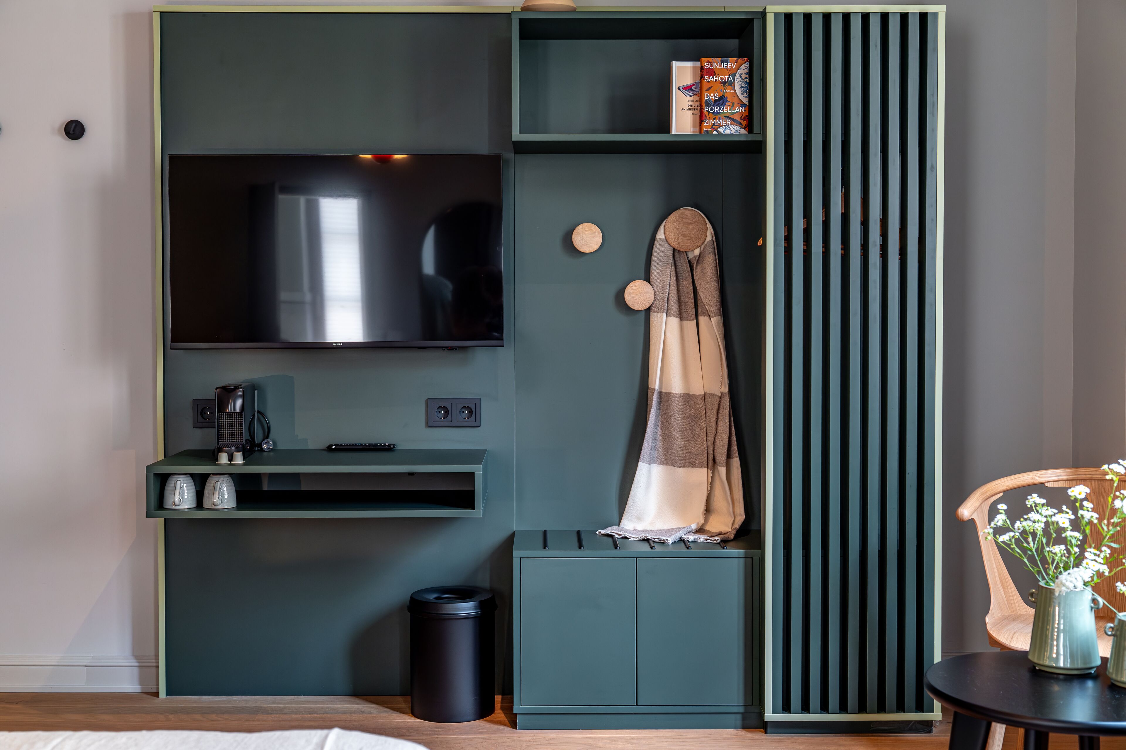 Una habitación moderna con televisión, un escritorio pequeño, un armario, un bote de basura y una silla con un florero.