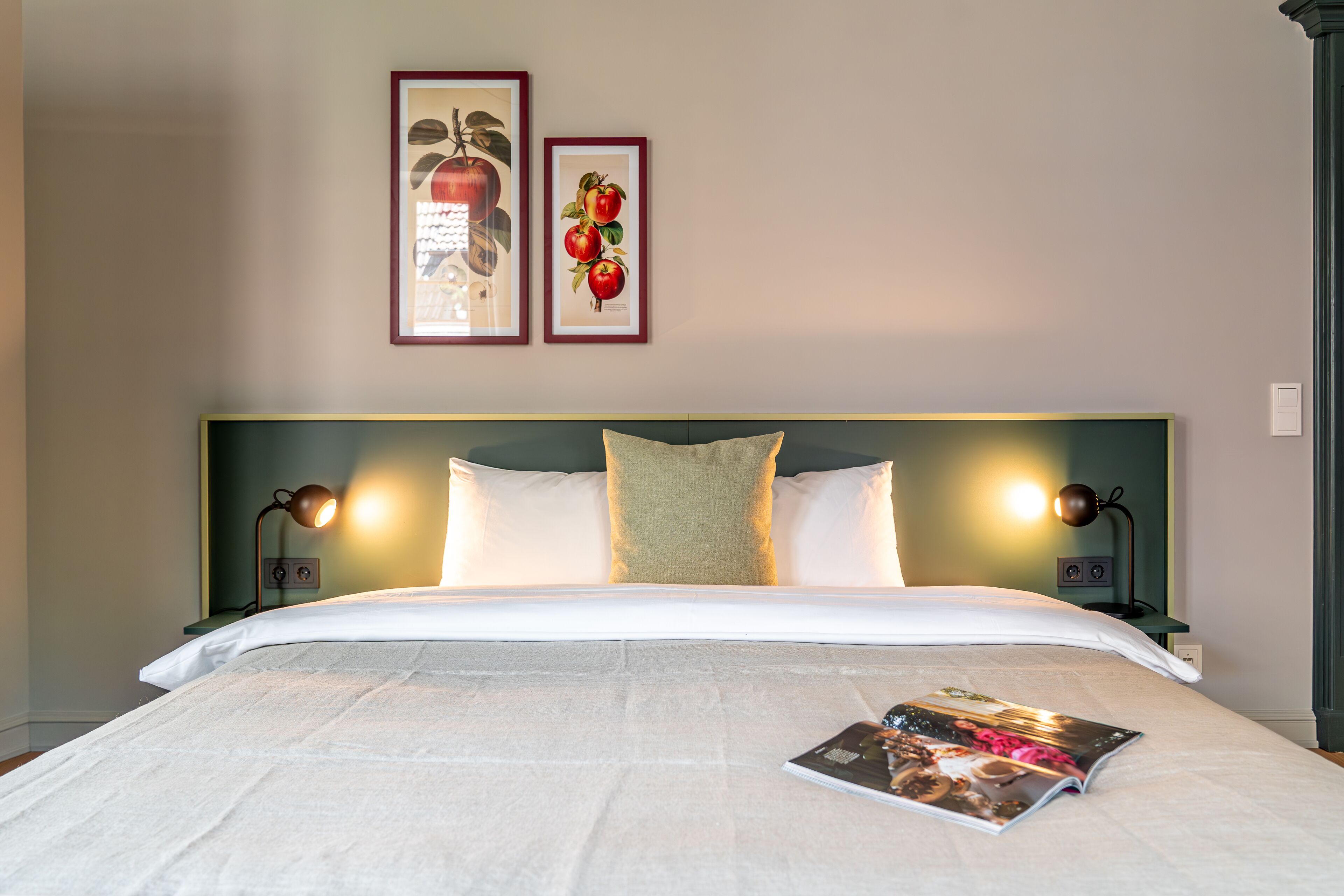 Een net opgemaakt bed met witte lakens, twee ingekaderde foto's aan de muur en een tijdschrift op het bed.