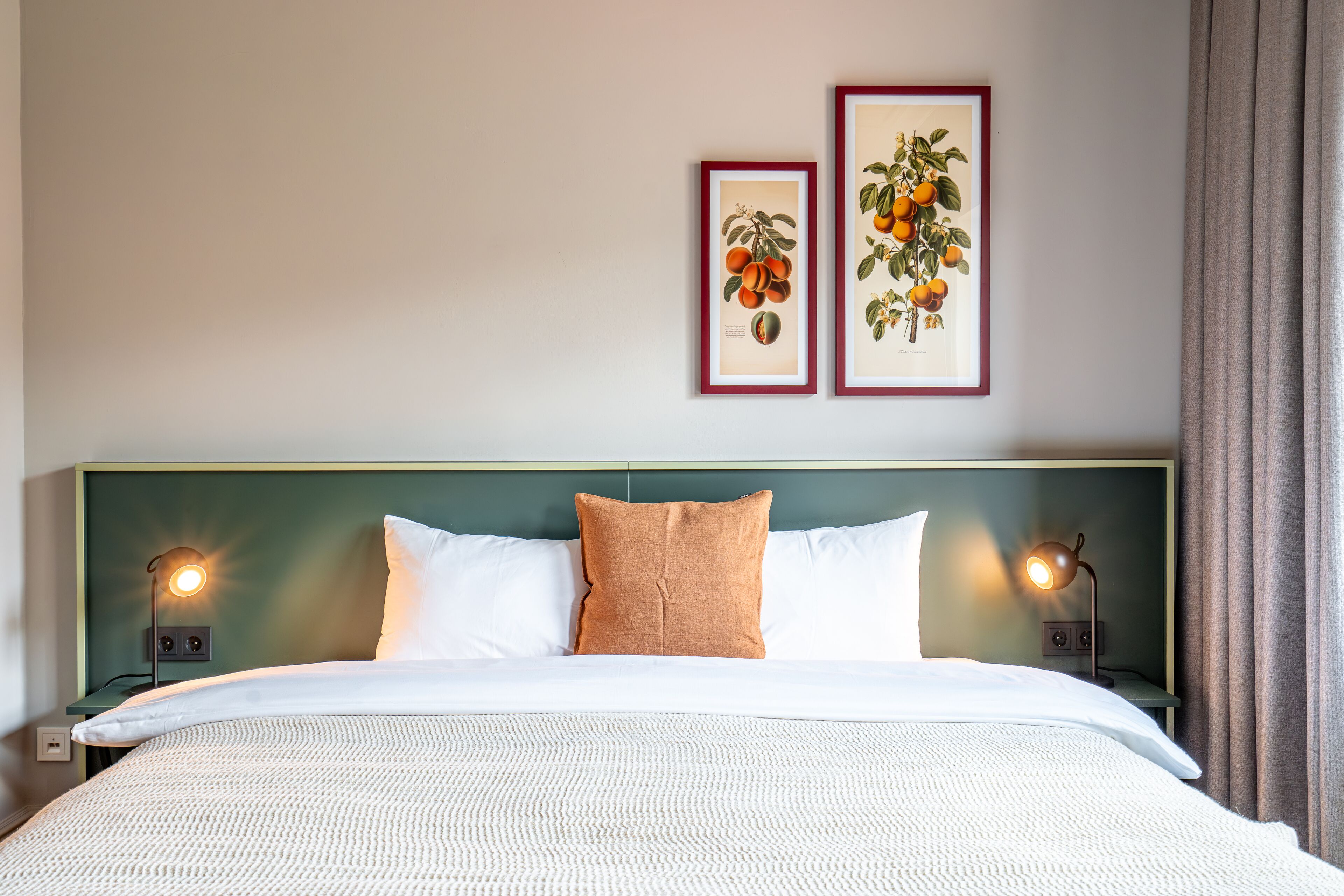 A neatly made bed with white linens, two pillows, and a green headboard. There are two framed botanical prints on the wall above the bed.