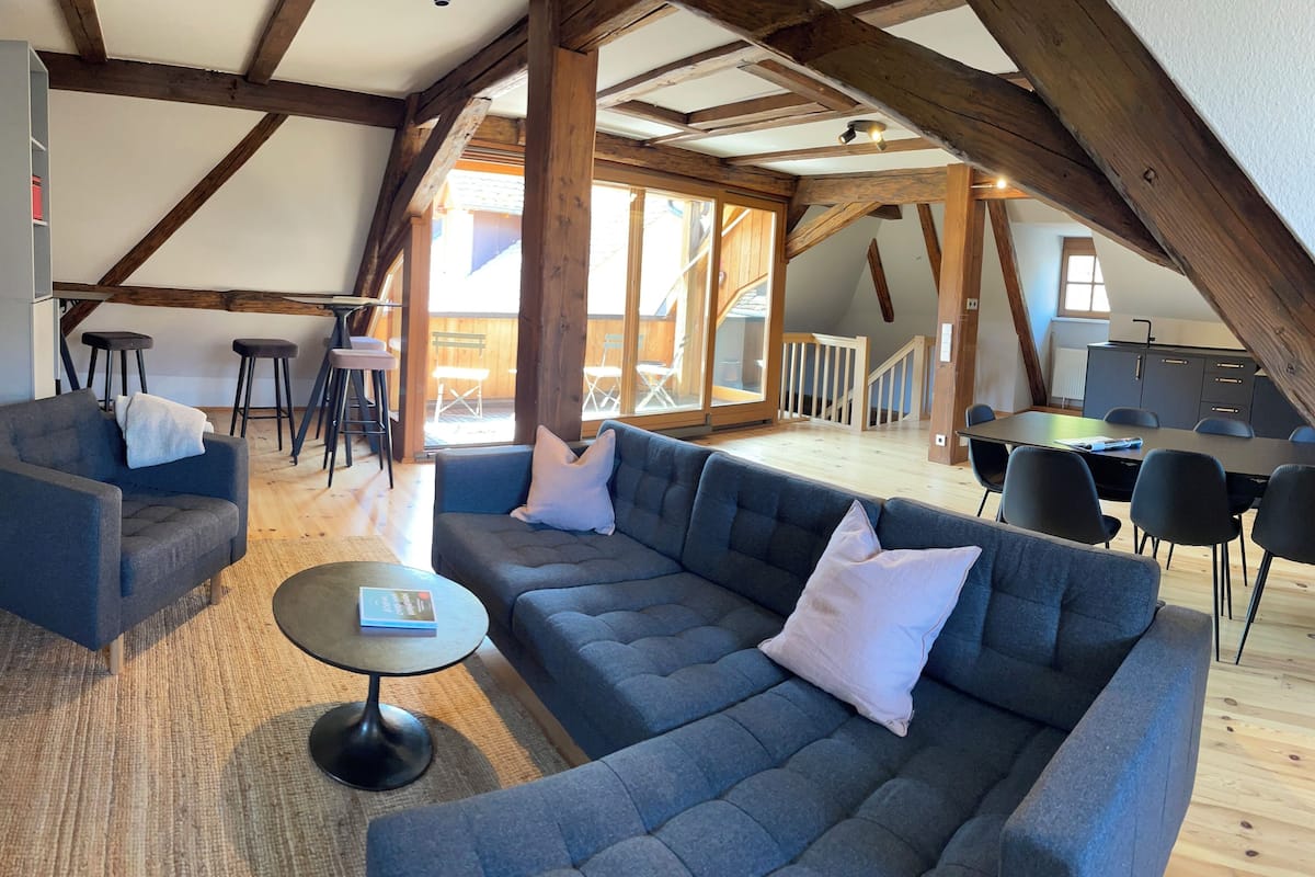 Un loft espacioso con techo alto, vigas de madera a la vista, un sofá modular, una pequeña mesa redonda y un comedor con sillas.