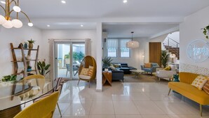 A modern living room with a glass dining table, yellow chairs, a sofa, and a hanging light fixture.