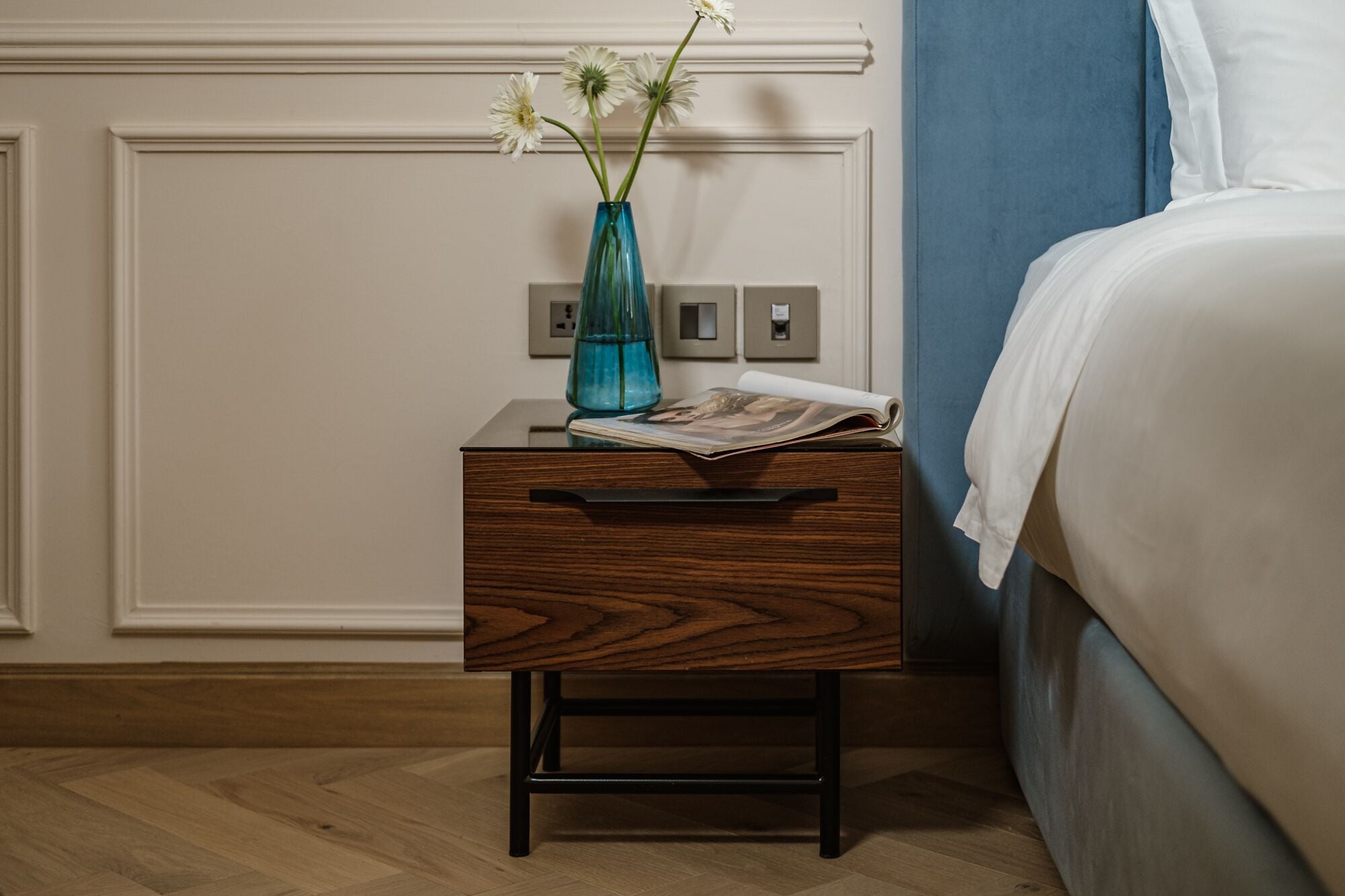 Une table de chevet en bois sur laquelle se trouve un vase bleu contenant des fleurs blanches, un magazine et une prise électrique.